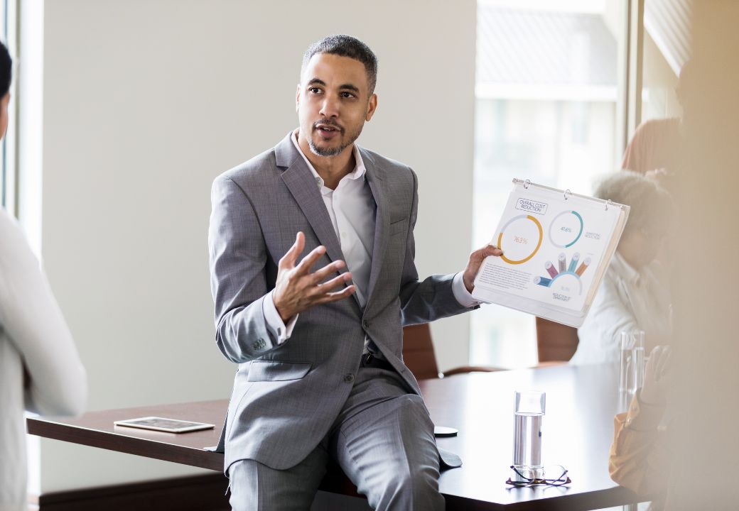 Business professional presenting data charts during a meeting while sitting on a conference table.