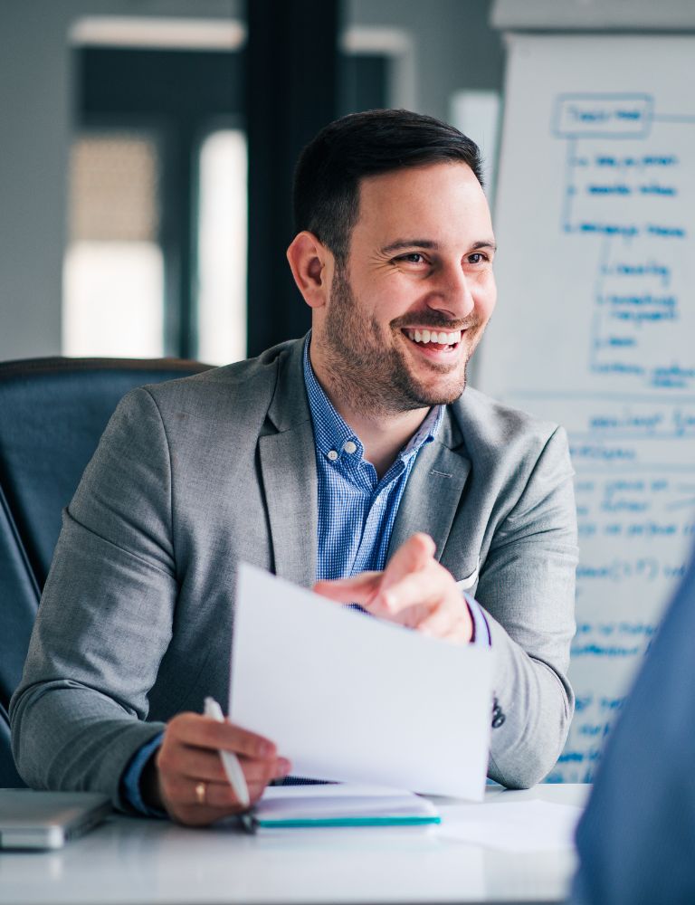 Smiling man in a gray suit, sitting at a desk with documents in hand, during a business meeting. Smiling man in a gray suit, sitting at a desk with documents in hand, during a business meeting.