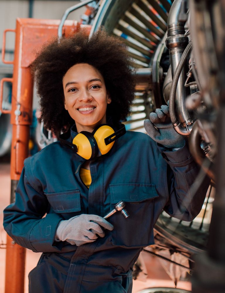 Vrouw in werkkleding met afro kapsel, lachend en staand bij een machine met een steeksleutel in de hand en gehoorbescherming om haar nek. Vrouw in werkkleding met afro kapsel, lachend en staand bij een machine met een steeksleutel in de hand en gehoorbescherming om haar nek.